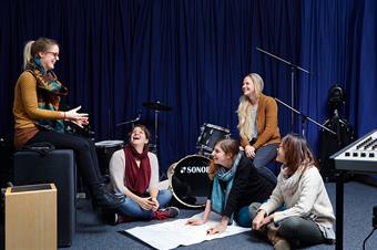 Dieses Bild zeigt eine Gruppe von weiblichen Studierenden umgeben von Instrumenten in einem Musikraum des Fachbereichs Sozial- und Kulturwissenschaften.